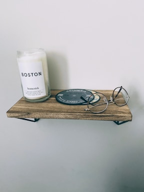 A minimalist wooden shelf is mounted on a plain wall. On the shelf, there is a large white candle in a glass jar labeled 'Boston' and 'homesick.' Next to it lies a round, dark coaster or record. A pair of round-framed glasses is placed beside it, adding to the simple decor.
