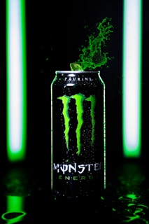 A can of Monster Energy drink is prominently featured, with green splashes erupting from its top. It is lit by vivid green lights, creating a dynamic and energetic atmosphere.