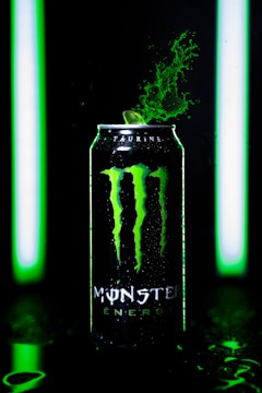 A can of Monster Energy drink is prominently featured, with green splashes erupting from its top. It is lit by vivid green lights, creating a dynamic and energetic atmosphere.