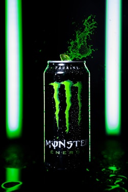 A can of Monster Energy drink is prominently featured, with green splashes erupting from its top. It is lit by vivid green lights, creating a dynamic and energetic atmosphere.