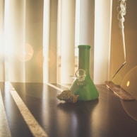 A green glass object resembling a bong sits on a dark surface. Beside it, a cannabis bud rests. Sunlight filters through window blinds, creating patterns of light and shadow across the table. To the right, incense is burning, with smoke drifting upward.