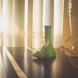 A green glass object resembling a bong sits on a dark surface. Beside it, a cannabis bud rests. Sunlight filters through window blinds, creating patterns of light and shadow across the table. To the right, incense is burning, with smoke drifting upward.