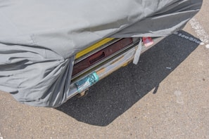 A partially covered vehicle with a gray tarp, revealing a yellow bumper and a license plate with recurring numbers. The scene is set on an asphalt surface with visible lines and markings.