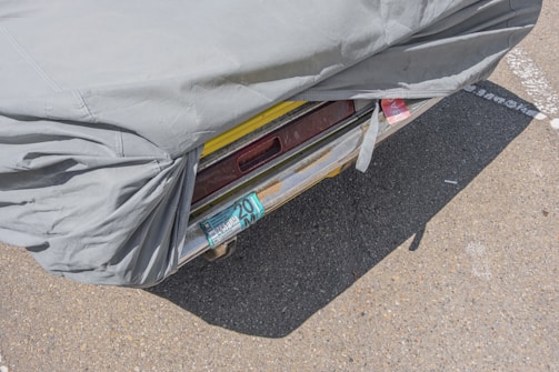 A partially covered vehicle with a gray tarp, revealing a yellow bumper and a license plate with recurring numbers. The scene is set on an asphalt surface with visible lines and markings.