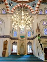 A beautifully ornate interior featuring Islamic architectural elements. There is an elaborate chandelier hanging from the ceiling with intricate patterns and Arabic calligraphy. The walls and arches are adorned with colorful stained glass windows and detailed mosaic tile work. A pulpit or minbar with detailed carvings is present on one side, and the soft, turquoise carpet covers the floor, leading to an area for prayer.