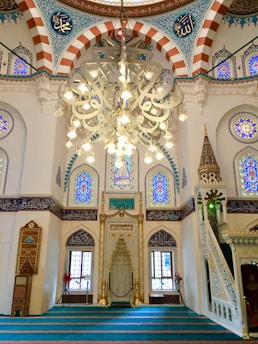 A beautifully ornate interior featuring Islamic architectural elements. There is an elaborate chandelier hanging from the ceiling with intricate patterns and Arabic calligraphy. The walls and arches are adorned with colorful stained glass windows and detailed mosaic tile work. A pulpit or minbar with detailed carvings is present on one side, and the soft, turquoise carpet covers the floor, leading to an area for prayer.
