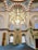 A beautifully ornate interior featuring Islamic architectural elements. There is an elaborate chandelier hanging from the ceiling with intricate patterns and Arabic calligraphy. The walls and arches are adorned with colorful stained glass windows and detailed mosaic tile work. A pulpit or minbar with detailed carvings is present on one side, and the soft, turquoise carpet covers the floor, leading to an area for prayer.