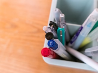A collection of pens and pencils is organized inside a light-colored container. The writing instruments feature varied designs and colors, including red, blue, and black, with some having a transparent casing.