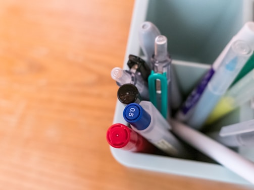 A collection of pens and pencils is organized inside a light-colored container. The writing instruments feature varied designs and colors, including red, blue, and black, with some having a transparent casing.