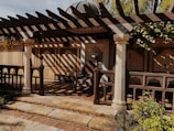 A rustic outdoor patio features a wooden pergola casting intricate shadows on the ground. The area is framed by decorative columns, and the backdrop consists of beige walls with floral motifs. There are plants and flowers in the foreground, creating a tranquil garden setting.