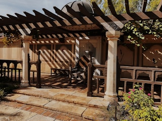 A rustic outdoor patio features a wooden pergola casting intricate shadows on the ground. The area is framed by decorative columns, and the backdrop consists of beige walls with floral motifs. There are plants and flowers in the foreground, creating a tranquil garden setting.