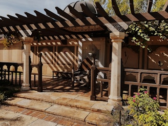 A rustic outdoor patio features a wooden pergola casting intricate shadows on the ground. The area is framed by decorative columns, and the backdrop consists of beige walls with floral motifs. There are plants and flowers in the foreground, creating a tranquil garden setting.