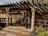 A rustic outdoor patio features a wooden pergola casting intricate shadows on the ground. The area is framed by decorative columns, and the backdrop consists of beige walls with floral motifs. There are plants and flowers in the foreground, creating a tranquil garden setting.