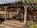 A rustic outdoor patio features a wooden pergola casting intricate shadows on the ground. The area is framed by decorative columns, and the backdrop consists of beige walls with floral motifs. There are plants and flowers in the foreground, creating a tranquil garden setting.