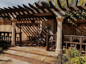 A rustic outdoor patio features a wooden pergola casting intricate shadows on the ground. The area is framed by decorative columns, and the backdrop consists of beige walls with floral motifs. There are plants and flowers in the foreground, creating a tranquil garden setting.