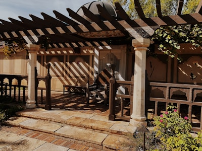 A rustic outdoor patio features a wooden pergola casting intricate shadows on the ground. The area is framed by decorative columns, and the backdrop consists of beige walls with floral motifs. There are plants and flowers in the foreground, creating a tranquil garden setting.