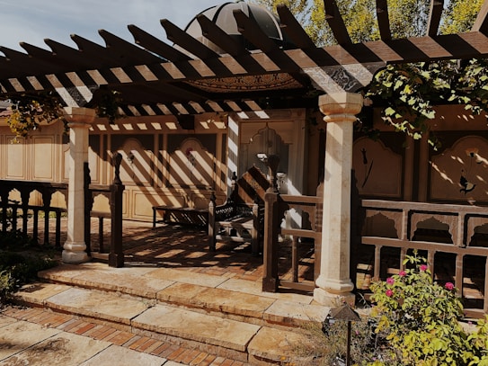 A rustic outdoor patio features a wooden pergola casting intricate shadows on the ground. The area is framed by decorative columns, and the backdrop consists of beige walls with floral motifs. There are plants and flowers in the foreground, creating a tranquil garden setting.