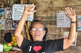 An elderly person is smiling broadly with both arms raised in a gesture of joy or celebration. They are wearing glasses and a black t-shirt with a heart symbol. In the background, there are woven bamboo walls decorated with various posters, menus, and fruit displayed on a counter.