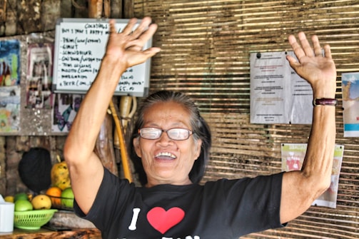 An elderly person is smiling broadly with both arms raised in a gesture of joy or celebration. They are wearing glasses and a black t-shirt with a heart symbol. In the background, there are woven bamboo walls decorated with various posters, menus, and fruit displayed on a counter.