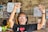 An elderly person is smiling broadly with both arms raised in a gesture of joy or celebration. They are wearing glasses and a black t-shirt with a heart symbol. In the background, there are woven bamboo walls decorated with various posters, menus, and fruit displayed on a counter.