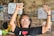 An elderly person is smiling broadly with both arms raised in a gesture of joy or celebration. They are wearing glasses and a black t-shirt with a heart symbol. In the background, there are woven bamboo walls decorated with various posters, menus, and fruit displayed on a counter.
