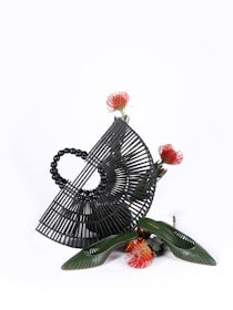 A black, semi-circular bamboo handbag with a structured lattice design is paired artistically with a pair of green shoes. Surrounding the items are vibrant red and orange protea flowers. The display is minimalistic on a white background, emphasizing the texture and form of the objects.