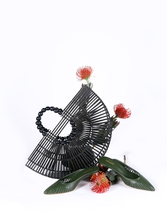 A black, semi-circular bamboo handbag with a structured lattice design is paired artistically with a pair of green shoes. Surrounding the items are vibrant red and orange protea flowers. The display is minimalistic on a white background, emphasizing the texture and form of the objects.