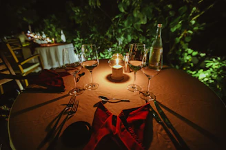 An intimate table set for a private dinner, glowing with candlelight and surrounded by dark wood and green plants.