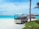 A small resort buggy is parked on a sandy path surrounded by greenery, with clear turquoise water and overwater bungalows in the background. The sky is bright and partly cloudy, and a palm tree is visible on the right side.