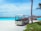 A small resort buggy is parked on a sandy path surrounded by greenery, with clear turquoise water and overwater bungalows in the background. The sky is bright and partly cloudy, and a palm tree is visible on the right side.