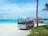 A small resort buggy is parked on a sandy path surrounded by greenery, with clear turquoise water and overwater bungalows in the background. The sky is bright and partly cloudy, and a palm tree is visible on the right side.