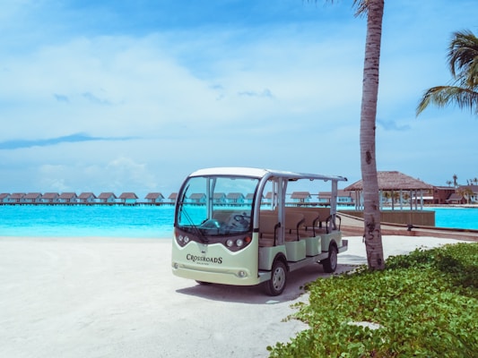 A small resort buggy is parked on a sandy path surrounded by greenery, with clear turquoise water and overwater bungalows in the background. The sky is bright and partly cloudy, and a palm tree is visible on the right side.