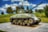 A World War II tank is displayed on a patterned concrete pedestal in a grassy area, surrounded by a black metal fence and trees. The tank is green, with visible identification marks including 'EXERCISE TIGER 749' and a star symbol. The sky is blue with patches of white clouds.