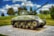 A World War II tank is displayed on a patterned concrete pedestal in a grassy area, surrounded by a black metal fence and trees. The tank is green, with visible identification marks including 'EXERCISE TIGER 749' and a star symbol. The sky is blue with patches of white clouds.