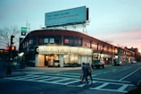A city street scene at dusk with a prominent brick building at the corner featuring a large sign for apartment rentals. The sidewalks are lined with bare trees and there are bright streetlights and store signs glowing. Several people can be seen walking across the crosswalk while a few cars are parked along the street. Above the building, a large billboard advertises cars, illuminated against the evening sky which transitions from blue to a soft pink.