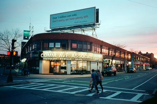 A city street scene at dusk with a prominent brick building at the corner featuring a large sign for apartment rentals. The sidewalks are lined with bare trees and there are bright streetlights and store signs glowing. Several people can be seen walking across the crosswalk while a few cars are parked along the street. Above the building, a large billboard advertises cars, illuminated against the evening sky which transitions from blue to a soft pink.
