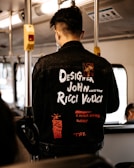 A person wearing a black denim jacket with various white and red text designs stands inside a bus. The jacket features words like 'Designer John Ricci Vucci' and phrases such as 'A Never Ending Journey'. The interior of the bus is visible, with metal poles and overhead lighting.