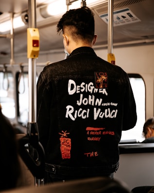 A person wearing a black denim jacket with various white and red text designs stands inside a bus. The jacket features words like 'Designer John Ricci Vucci' and phrases such as 'A Never Ending Journey'. The interior of the bus is visible, with metal poles and overhead lighting.
