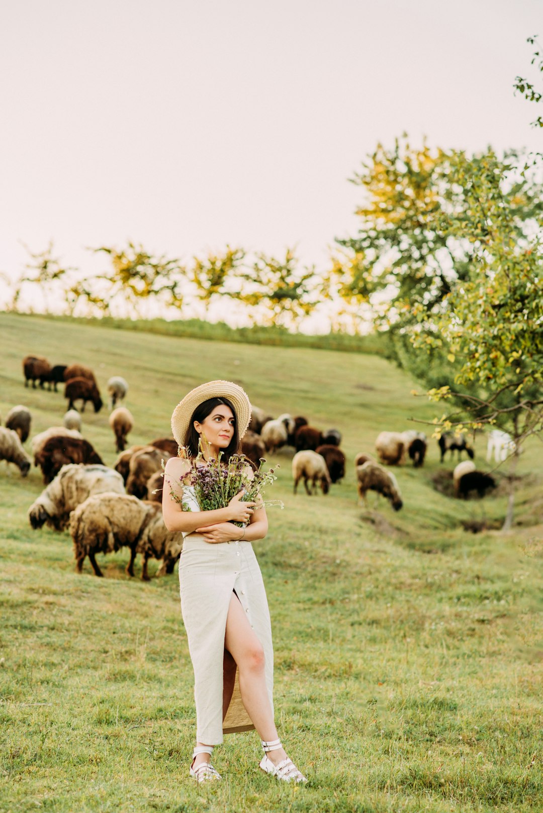 woman in white pants standing on green grass field with sheep during daytime