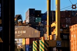 Stacks of shipping containers in various colors are piled high at a port. A large crane is actively lifting one container, which prominently displays the letters 'CMA CGM'. The sun casts shadows and a warm glow, creating a busy yet calm industrial scene.