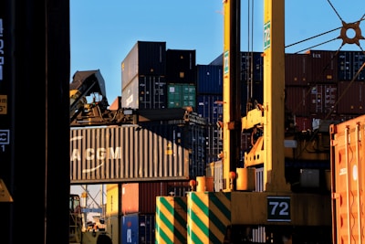 Stacks of shipping containers in various colors are piled high at a port. A large crane is actively lifting one container, which prominently displays the letters 'CMA CGM'. The sun casts shadows and a warm glow, creating a busy yet calm industrial scene.