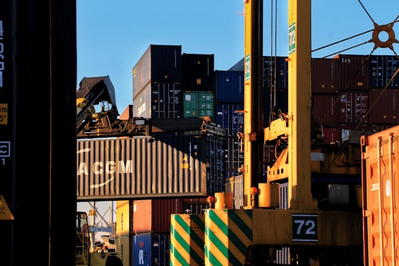 Stacks of shipping containers in various colors are piled high at a port. A large crane is actively lifting one container, which prominently displays the letters 'CMA CGM'. The sun casts shadows and a warm glow, creating a busy yet calm industrial scene.