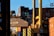 Stacks of shipping containers in various colors are piled high at a port. A large crane is actively lifting one container, which prominently displays the letters 'CMA CGM'. The sun casts shadows and a warm glow, creating a busy yet calm industrial scene.