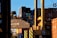 Stacks of shipping containers in various colors are piled high at a port. A large crane is actively lifting one container, which prominently displays the letters 'CMA CGM'. The sun casts shadows and a warm glow, creating a busy yet calm industrial scene.