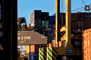 Stacks of shipping containers in various colors are piled high at a port. A large crane is actively lifting one container, which prominently displays the letters 'CMA CGM'. The sun casts shadows and a warm glow, creating a busy yet calm industrial scene.