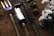 A set of garden tools including a fork, a spade, and a trowel are laid out on a rustic wooden surface. Nearby, there are pine cones and a bunch of white cotton flowers, adding a natural and earthy feel to the composition.