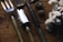 A set of garden tools including a fork, a spade, and a trowel are laid out on a rustic wooden surface. Nearby, there are pine cones and a bunch of white cotton flowers, adding a natural and earthy feel to the composition.