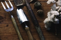 A set of garden tools including a fork, a spade, and a trowel are laid out on a rustic wooden surface. Nearby, there are pine cones and a bunch of white cotton flowers, adding a natural and earthy feel to the composition.