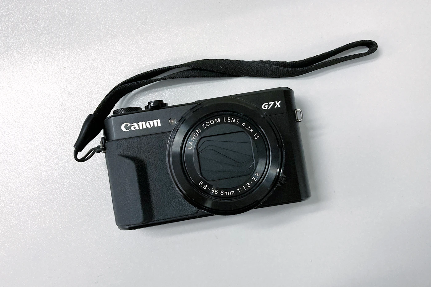 A small black pocket compact camera with a built-in zoom lens, lying on a white surface with its wrist strap