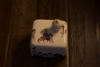 A small, square white ceramic oil lamp placed on a wooden surface. The lamp features intricate illustrations of people and animals in traditional attire, one person is riding an animal while others appear to be walking alongside. The wooden surface conveys a rustic look, and the lighting is dim.